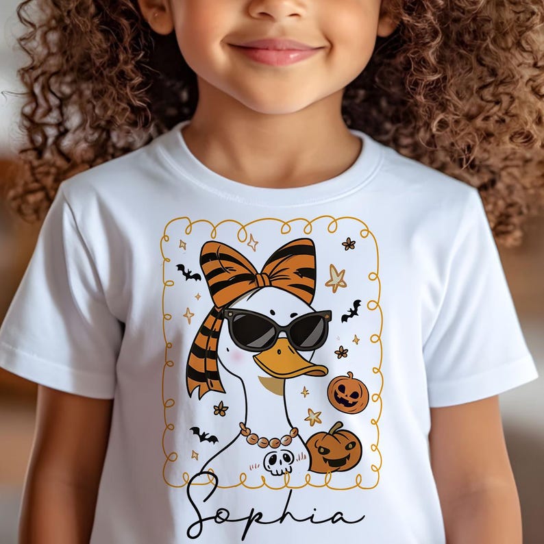 Op de afbeelding: Wit T-shirt met een cartoon eend met een zonnebril, een strik en een ketting. De eend is omringd door Halloween-thema graphics, waaronder pompoenen en vleermuizen. De naam "Sophia" staat eronder in cursief.