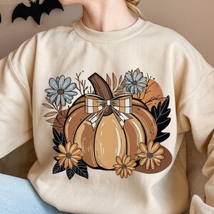 Puede incluir: Sudadera color crema con un gran diseño de calabaza con lazo a cuadros, rodeada de flores y hojas en tonos marrón, naranja y azul. El diseño tiene un tema otoñal.