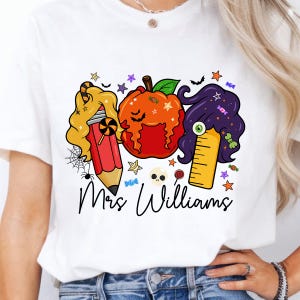 May include: White t-shirt featuring a Halloween-themed graphic. The design includes a pencil, pumpkin, and ruler with hair, along with the text "Mrs. Williams". The graphic is decorated with stars, candy, and a spider web.