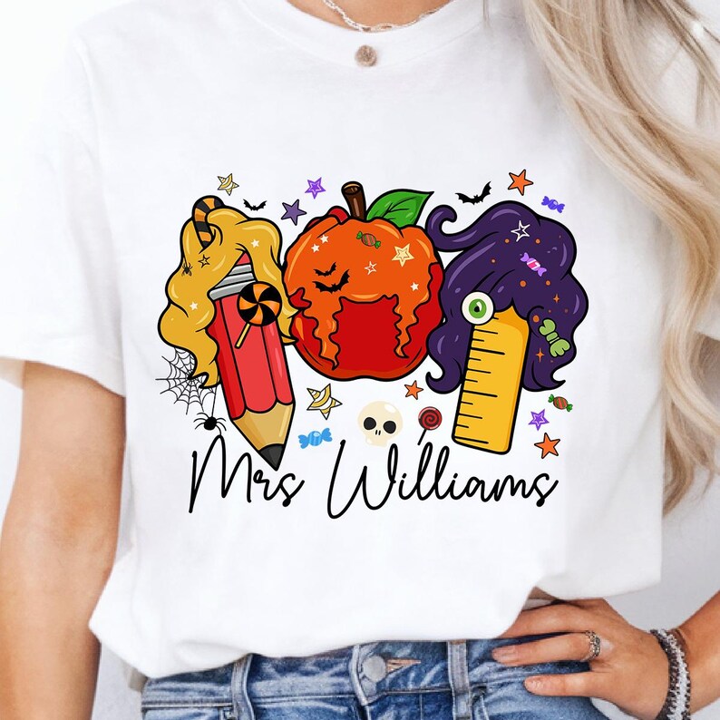 May include: White t-shirt featuring a Halloween-themed graphic. The design includes a pencil, pumpkin, and ruler with hair, along with the text "Mrs. Williams". The graphic is decorated with stars, candy, and a spider web.