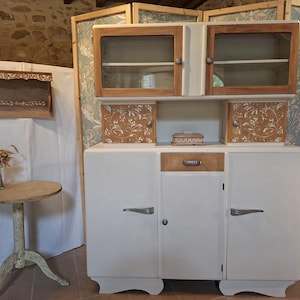 Peut inclure: Un buffet vintage blanc et bois avec des armoires supérieures vitrées et des motifs floraux décoratifs. Le buffet a un tiroir central et deux portes inférieures avec des poignées argentées. Une petite table ronde et un paravent décoratif sont également visibles.