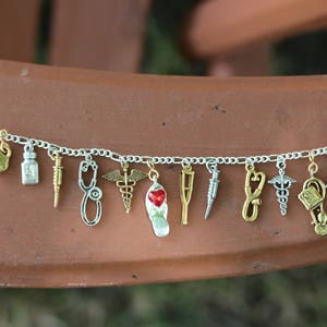 Könnte beinhalten: Ein silbernes Armband mit verschiedenen medizinischen Charms, darunter ein Stethoskop, ein Caduceus, ein Herz, eine Krücke, eine Spritze, eine Pillenflasche, ein Erste-Hilfe-Kasten und ein Krankenwagen.
