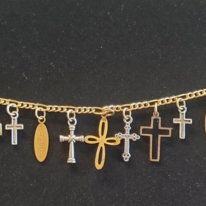 May include: Gold-tone chain bracelet featuring various cross charms in silver and gold. Some crosses have intricate designs, while others are simple. An oval charm with text is also included.