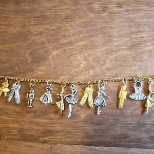 Pode incluir: Um bracelete de corrente dourada com vários pingentes, incluindo bailarinas, um quebra-nozes e uma sapatilha de balé. Os pingentes são prateados e dourados.
