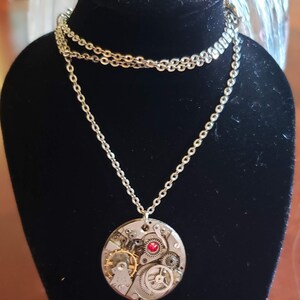 Puede incluir: Un collar de cadena de plata con un colgante redondo que presenta un diseño de mecanismo de relojería detallado con un acento de joya roja.