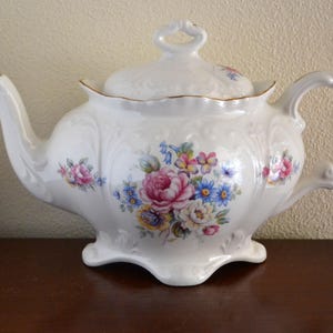 以下が含まれることがあります： 花柄の白い磁器製のティーポット。ティーポットの蓋と注ぎ口の周りに金色の縁取りが施されています。花はピンク、ブルー、イエローです。