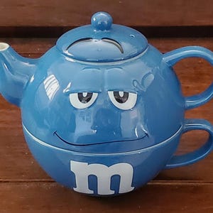 Puede incluir: Tetera de cerámica azul con una cara de dibujos animados y el logotipo de M&M's. La tetera tiene una tapa y un asa en el lateral. También tiene una taza separada con asa.