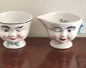 Puede incluir: Dos tazas de cerámica blanca con caras. Una taza tiene una cara guiñando un ojo y dice "Bailey's" en el lateral. La otra taza tiene un ojo cerrado y dice "Yum" y "Bailey's" en el lateral.