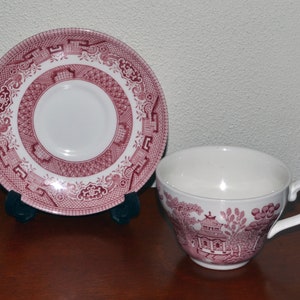 May include: A pink and white teacup and saucer set featuring a traditional Chinese design. The saucer has a detailed pattern of a pagoda and flowers. The teacup has a matching design on the side.