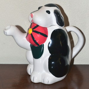 May include: A white and black ceramic teapot shaped like a dog holding a red flower with a yellow center. The teapot has a spout and a handle.