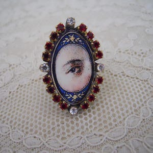 May include: Oval-shaped brooch with a painted eye design. The brooch features a red and clear gemstone border, a blue and gold inner border, and a central image of an eye. The brooch is set against a white lace background.