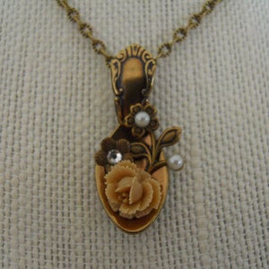 May include: A gold spoon necklace with a floral design. The spoon is decorated with a white rose, white pearl accents, and a small rhinestone.