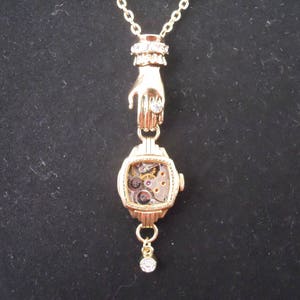 May include: A gold necklace with a hand pendant holding a small, open-faced watch with visible gears. The watch has a clear face and a gold chain attached to the bottom.