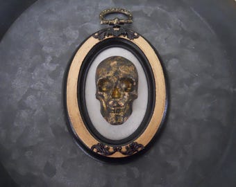 May include: A gold skull is displayed in a black and gold oval frame with a decorative gold clasp at the top. The frame is set against a dark gray background.
