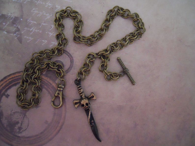 Pode incluir: Um colar de corrente dourada com um pingente de caveira e espada. A corrente &eacute; uma corrente de cabo duplo com um fecho de barra em T. O pingente &eacute; uma caveira detalhada com uma espada que a atravessa.