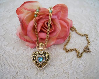 May include: A gold-toned heart-shaped pendant necklace with a blue gemstone center and clear crystal accents. The pendant hangs from a delicate chain. The necklace is set against a pink rose and a white lace background.