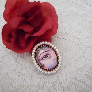 May include: An oval-shaped brooch featuring an eye design, framed with pearls and red beads. A large red rose rests beside the brooch on a white lace surface. The brooch is a unique accessory.