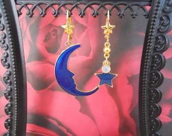 May include: Gold-colored earrings with celestial designs. One earring features a blue crescent moon with a face, the other has a blue star pendant with a smaller star and beads. The earrings hang from a gold star.