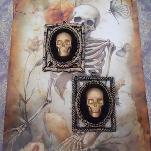 May include: Two framed skull pendants with gold-colored skulls, set against a backdrop of a skeleton, flowers, and butterflies. The frames are dark with gold accents. The background has a vintage, antique aesthetic.