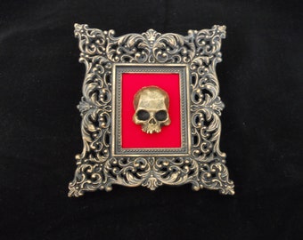 May include: A gold skull sculpture displayed in a ornate gold frame with a red velvet background.