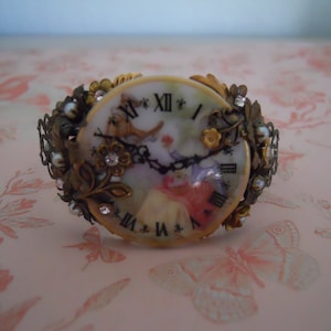 May include: A vintage-style clock face with Roman numerals and a floral design, set in a gold-toned metal frame with rhinestones and pearls.
