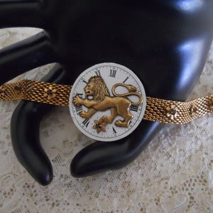 May include: A gold-tone bracelet with a white clock face featuring a lion and Roman numerals. The bracelet is made of a mesh design with small black flower accents.