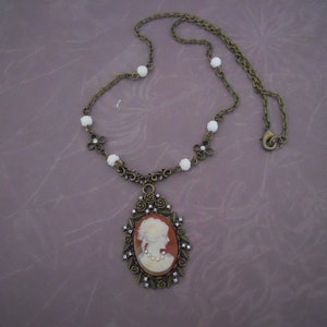 May include: A gold tone necklace with a cameo pendant featuring a white profile of a woman. The necklace has a chain with white beads and a lobster claw clasp.