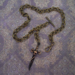 Pode incluir: Um colar de corrente de ouro com um pingente de caveira e espada. A corrente &eacute; grossa, de estilo tran&ccedil;ado e tem um fecho de trava. O pingente &eacute; uma caveira detalhada com uma espada atravessando-a.
