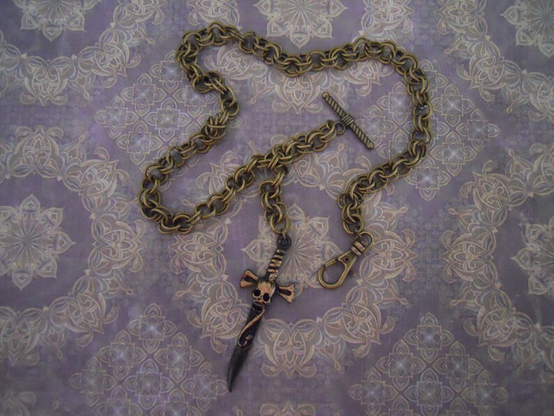 Pode incluir: Um colar de corrente de ouro com um pingente de caveira e espada. A corrente &eacute; grossa, de estilo tran&ccedil;ado e tem um fecho de trava. O pingente &eacute; uma caveira detalhada com uma espada atravessando-a.