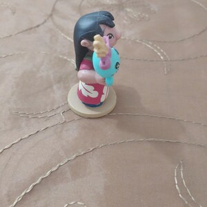 May include: A figurine of a character with black hair, wearing a red and white dress, holding a blue creature. The figurine is on a round, light-colored base. The character's face is visible, and the figurine is set against a neutral background.