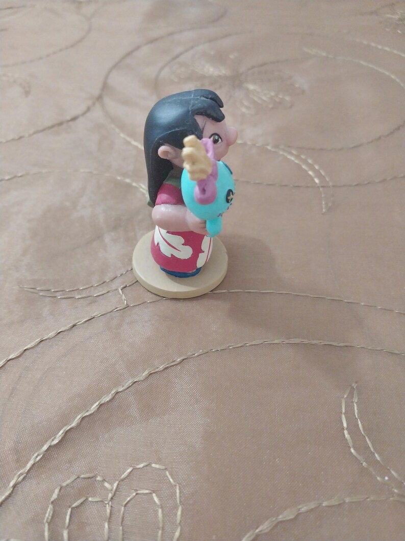 May include: A figurine of a character with black hair, wearing a red and white dress, holding a blue creature. The figurine is on a round, light-colored base. The character's face is visible, and the figurine is set against a neutral background.