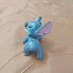 May include: A blue Stitch figurine with large ears, a wide mouth, and large eyes. The figurine is in a playful pose, with its back to the viewer. The ears are pink on the inside. The figurine is a collectible item.