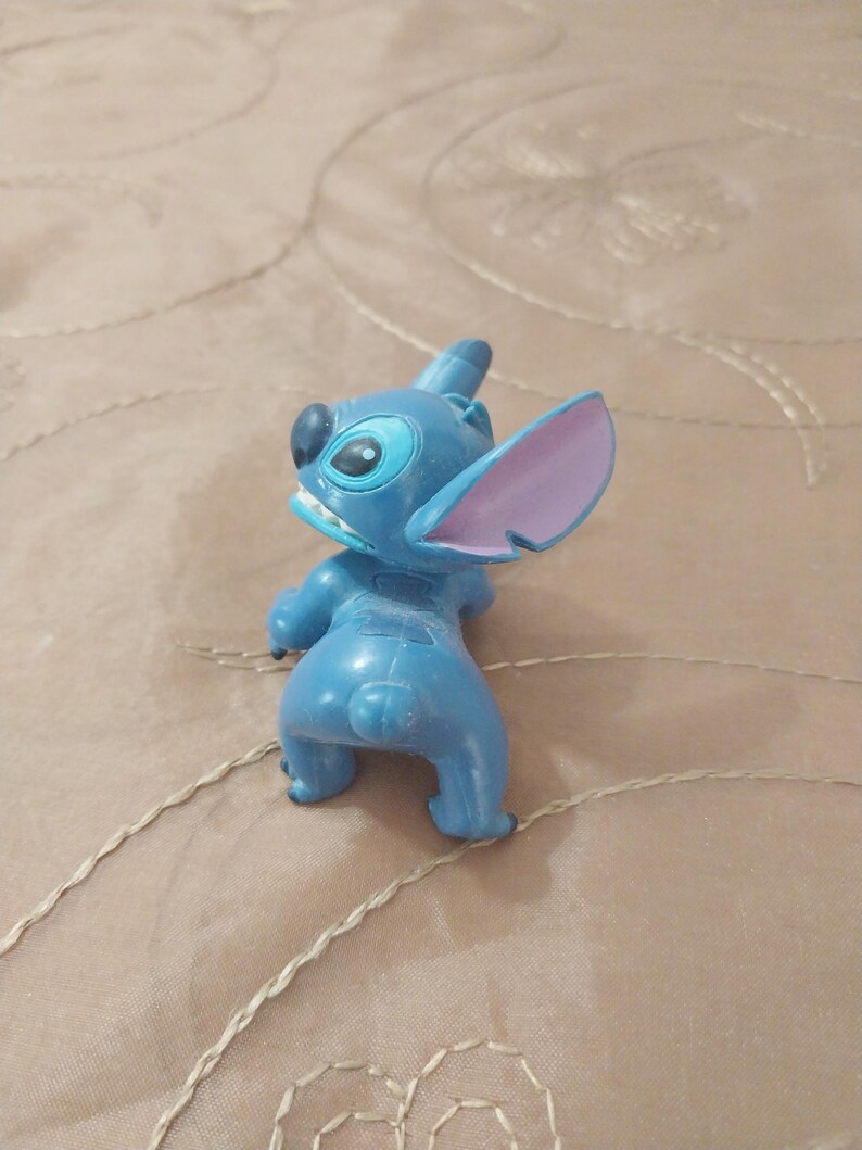 May include: A blue Stitch figurine with large ears, a wide mouth, and large eyes. The figurine is in a playful pose, with its back to the viewer. The ears are pink on the inside. The figurine is a collectible item.