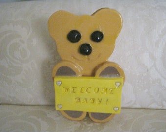 May include: A yellow teddy bear holding a sign that says "Welcome Baby!" The sign has white hearts on the corners.
