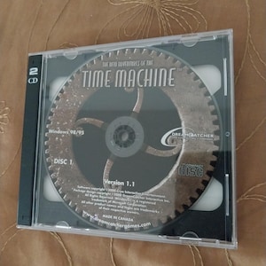 May include: A CD case for the computer game "The New Adventures of the Time Machine". The case is black with a clear plastic insert showing the CD. The CD has a metallic design with the game title and logo. The case also includes the game's version number, copyright information, and the Dreamcatcher Interactive logo.