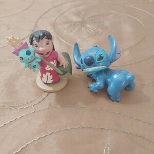 May include: Two figurines from the animated film Lilo & Stitch. One figurine depicts a girl in a red dress holding a doll. The other is a blue alien character with large ears and a toothy grin.