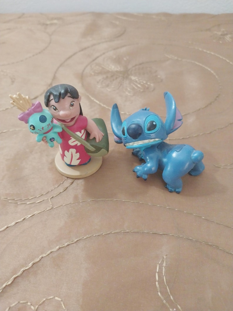May include: Two figurines from the animated film Lilo & Stitch. One figurine depicts a girl in a red dress holding a doll. The other is a blue alien character with large ears and a toothy grin.