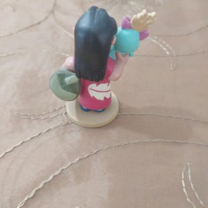 May include: A figurine of a character from the animated film Lilo & Stitch. The figure is wearing a pink dress and holding a teal object with a pink bow. The character has black hair and is standing on a beige base.