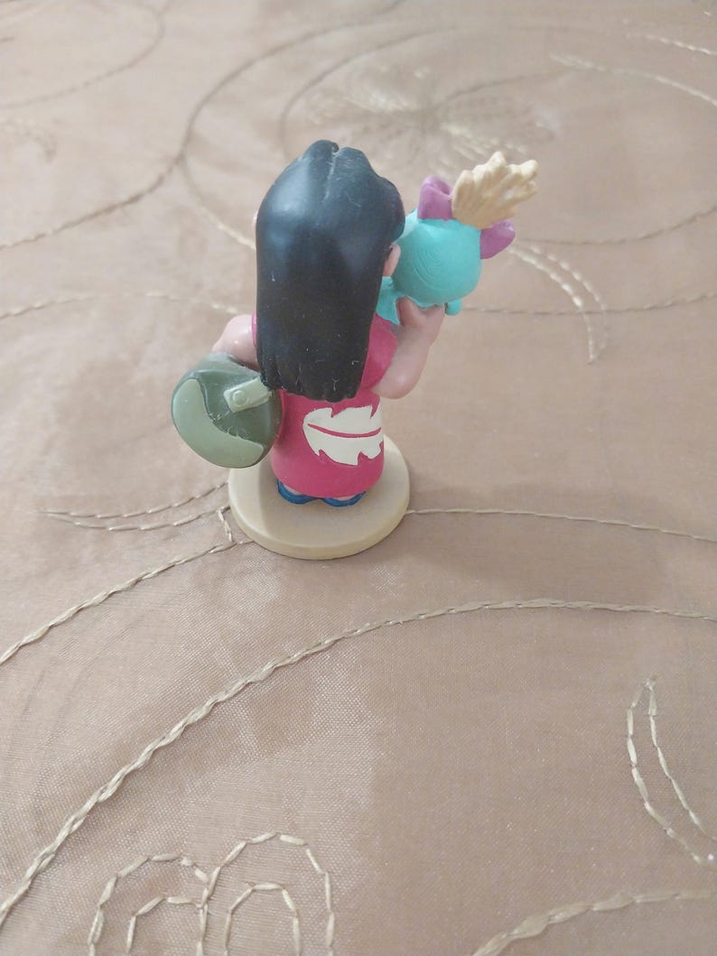 May include: A figurine of a character from the animated film Lilo & Stitch. The figure is wearing a pink dress and holding a teal object with a pink bow. The character has black hair and is standing on a beige base.