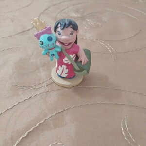 May include: A figurine of Lilo Pelekai from the Disney movie Lilo & Stitch. Lilo is wearing a red dress with white floral designs and carrying a green bag. She is holding a blue Stitch doll with a pink bow.