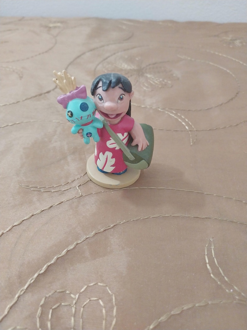 May include: A figurine of Lilo Pelekai from the Disney movie Lilo & Stitch. Lilo is wearing a red dress with white floral designs and carrying a green bag. She is holding a blue Stitch doll with a pink bow.