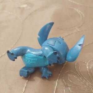 May include: A blue Stitch figurine from Disney. The figurine is in a crawling position, with large ears and a distinctive head shape. The underside of the figurine has the text "&copy; Disney China".