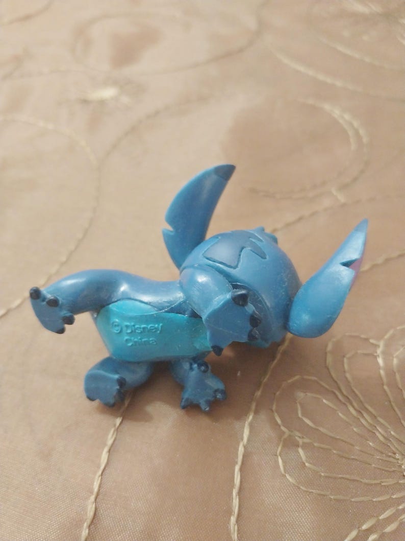 May include: A blue Stitch figurine from Disney. The figurine is in a crawling position, with large ears and a distinctive head shape. The underside of the figurine has the text "&copy; Disney China".