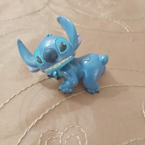 May include: A blue Stitch figurine, a character from the Disney movie Lilo & Stitch, is shown in a playful pose. The figurine has large ears, a wide grin, and a detailed design. The figure is a collectible item.