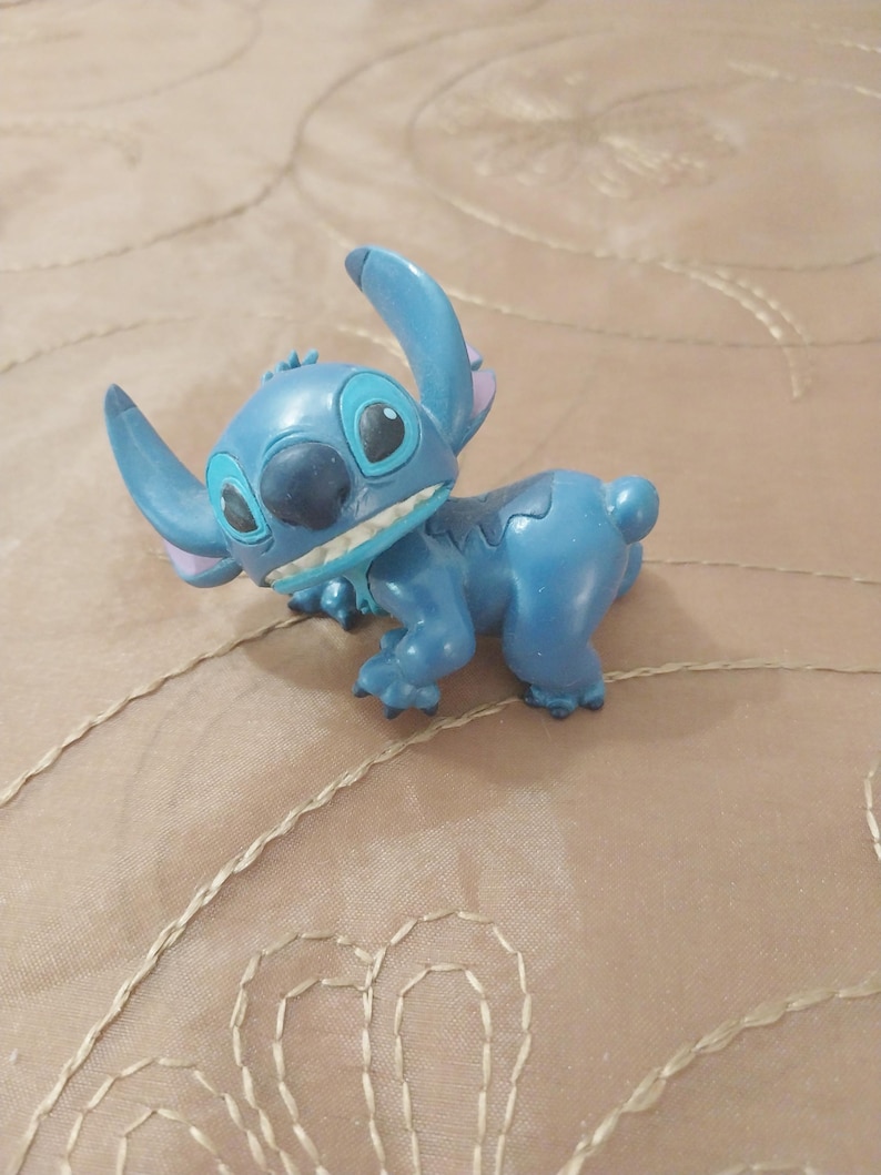 May include: A blue Stitch figurine, a character from the Disney movie Lilo & Stitch, is shown in a playful pose. The figurine has large ears, a wide grin, and a detailed design. The figure is a collectible item.