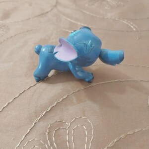 May include: A blue figurine of the character Stitch from the animated film Lilo & Stitch. The figurine has large ears, a small tail, and is in a walking pose. The ears are a light purple color.