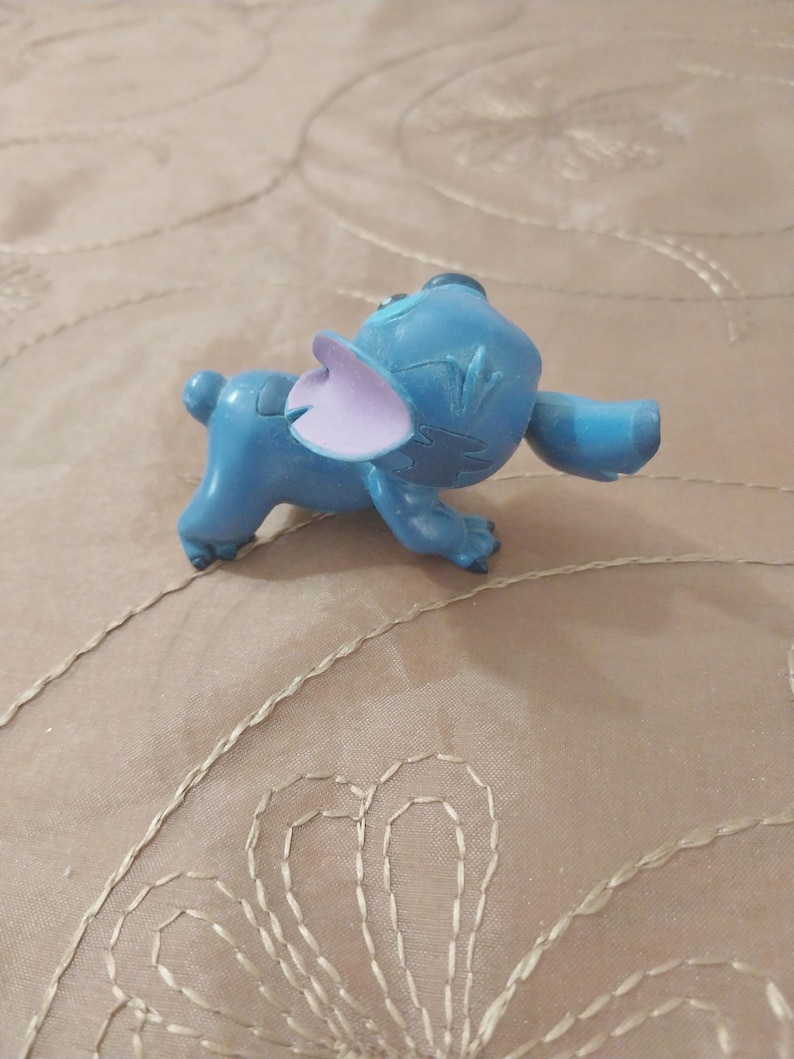 May include: A blue figurine of the character Stitch from the animated film Lilo & Stitch. The figurine has large ears, a small tail, and is in a walking pose. The ears are a light purple color.