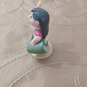 May include: A small figurine of a cartoon character, featuring black hair, a pink shirt, and a green skirt. The figurine is on a white base and has a small, yellow horn-like detail on its head. The figure is in a seated position.