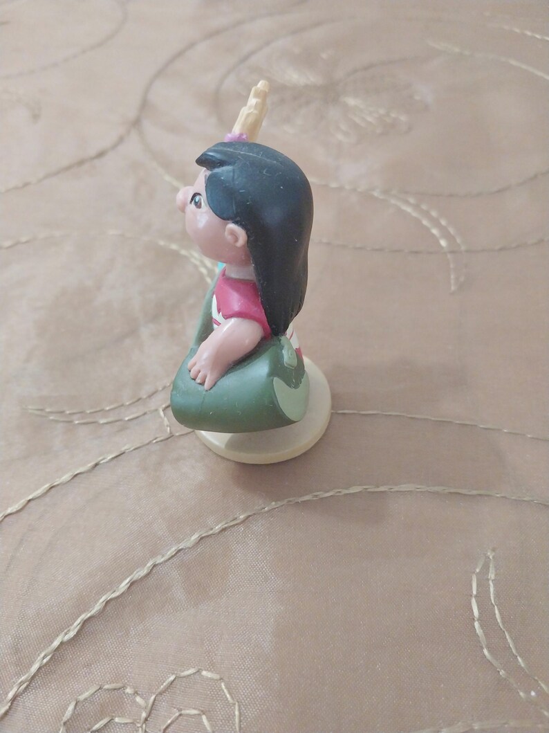 May include: A small figurine of a cartoon character, featuring black hair, a pink shirt, and a green skirt. The figurine is on a white base and has a small, yellow horn-like detail on its head. The figure is in a seated position.