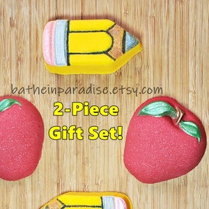 May include: A 2-piece gift set featuring bath bombs shaped like a yellow pencil and a red apple. The pencil bath bombs have a pink eraser and a silver tip. The apple bath bombs have green leaves and stems. The text "2-Piece Gift Set!" is visible.